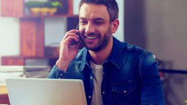 Hombre hablando por teléfono y sonriendo frente a una computadora portátil.