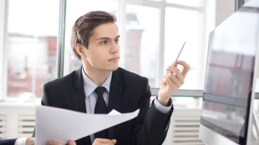 Hombre en traje revisa documentos frente a una pantalla de computadora.