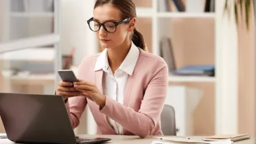 Mujer con gafas trabajando en una oficina mientras usa su teléfono móvil.