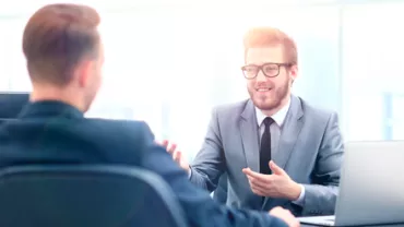 Dos hombres en traje, uno hablando y otro escuchando, en una oficina.