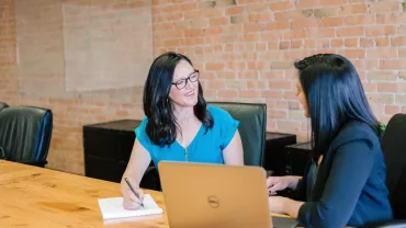 Dos mujeres conversan sonrientes en una sala de reuniones.