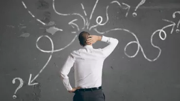 Persona pensativa ante una pizarra con signos de interrogación y flechas.