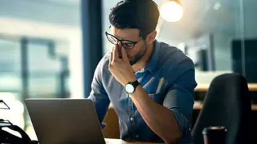 Hombre con gafas frente a una laptop, parece pensativo.
