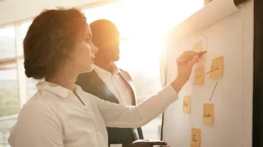 Dos personas discuten ideas en un pizarrón con luz de fondo.
