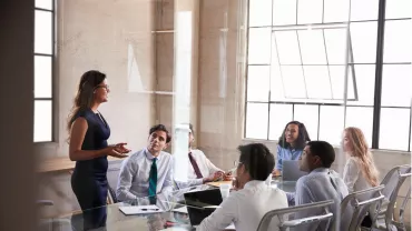 Una mujer presenta en una reunión de negocios a cinco colegas en una sala con ventanas grandes.