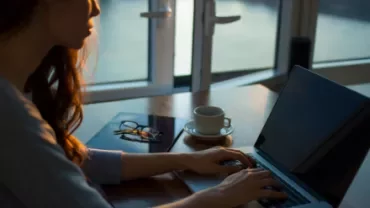 Mujer trabajando en laptop cerca de una ventana al atardecer.