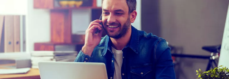 Hombre hablando por teléfono y sonriendo frente a una computadora portátil.