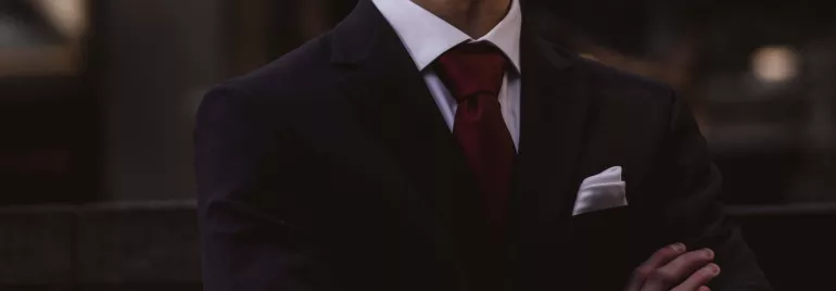 Hombre con traje oscuro y corbata roja cruzado de brazos en exterior.
