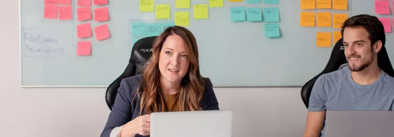 Dos personas trabajando en portátiles, con notas adhesivas en la pizarra.