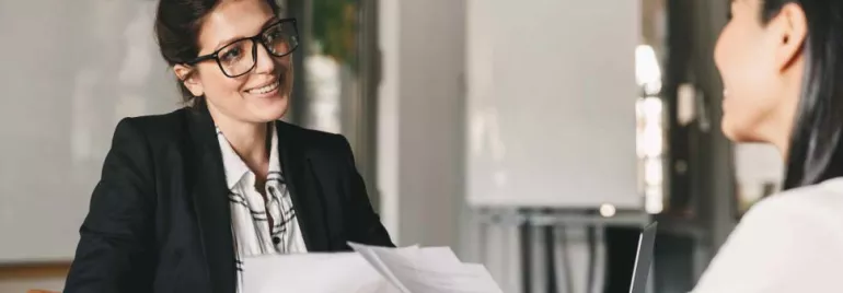 Dos mujeres en una reunión de negocios, una hablando y otra escuchando.