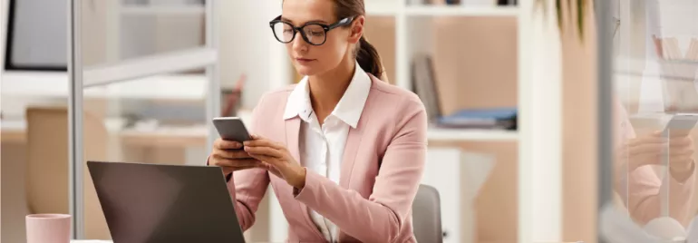 Mujer con gafas trabajando en una oficina mientras usa su teléfono móvil.