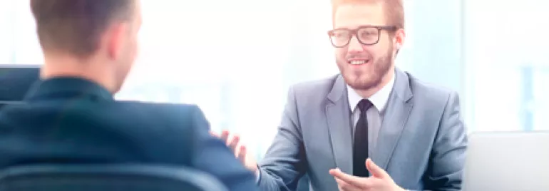 Dos hombres en traje, uno hablando y otro escuchando, en una oficina.