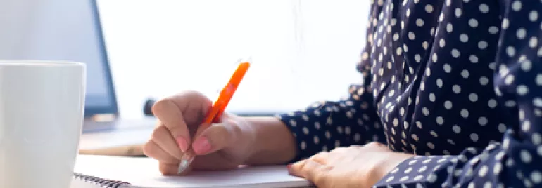Persona escribiendo en un cuaderno con una taza y una laptop enfrente.