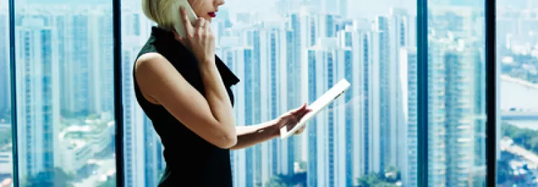 Mujer hablando por teléfono y usando una tableta frente a una ventana con vista a la ciudad.