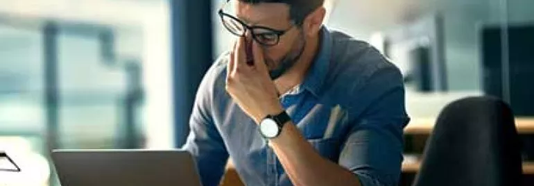 Hombre con gafas frente a una laptop, parece pensativo.