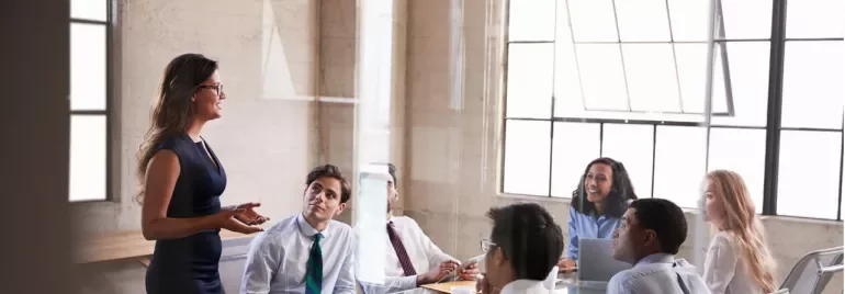 Una mujer presenta en una reunión de negocios a cinco colegas en una sala con ventanas grandes.