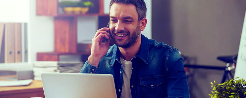 Hombre hablando por teléfono y sonriendo frente a una computadora portátil.
