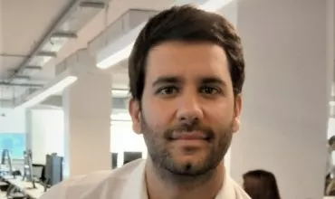 Hombre sonriente en una oficina moderna con fondo de escritorios y luces.