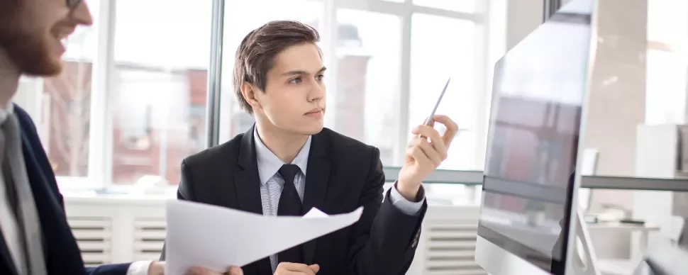 Hombre en traje revisa documentos frente a una pantalla de computadora.