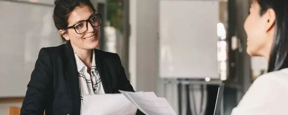Dos mujeres en una reunión de negocios, una hablando y otra escuchando.