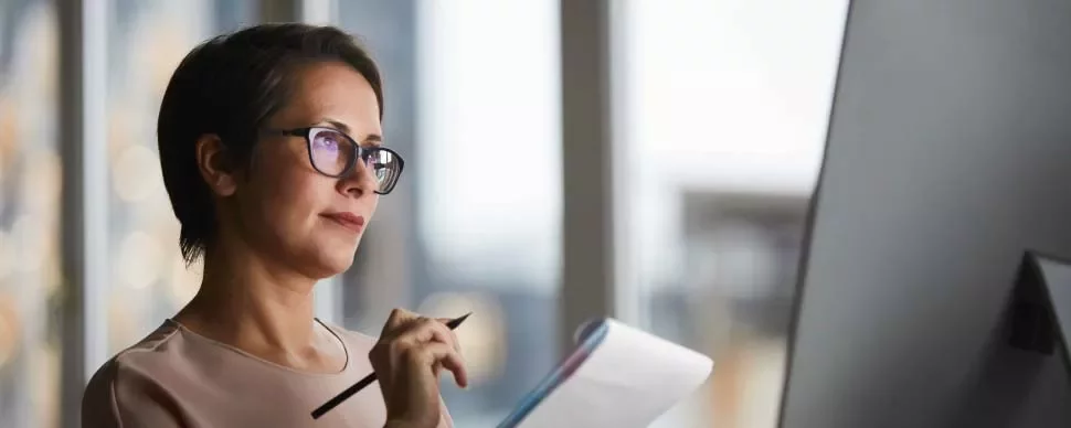 Mujer concentrada con gafas, sosteniendo un lápiz y libreta, mirando una pantalla.