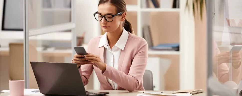 Mujer con gafas trabajando en una oficina mientras usa su teléfono móvil.