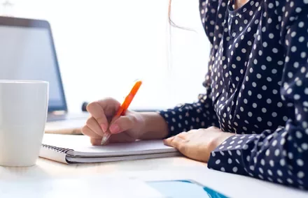 Persona escribiendo en un cuaderno con una taza y una laptop enfrente.
