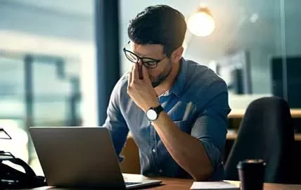 Hombre con gafas frente a una laptop, parece pensativo.
