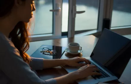 Mujer trabajando en laptop cerca de una ventana al atardecer.